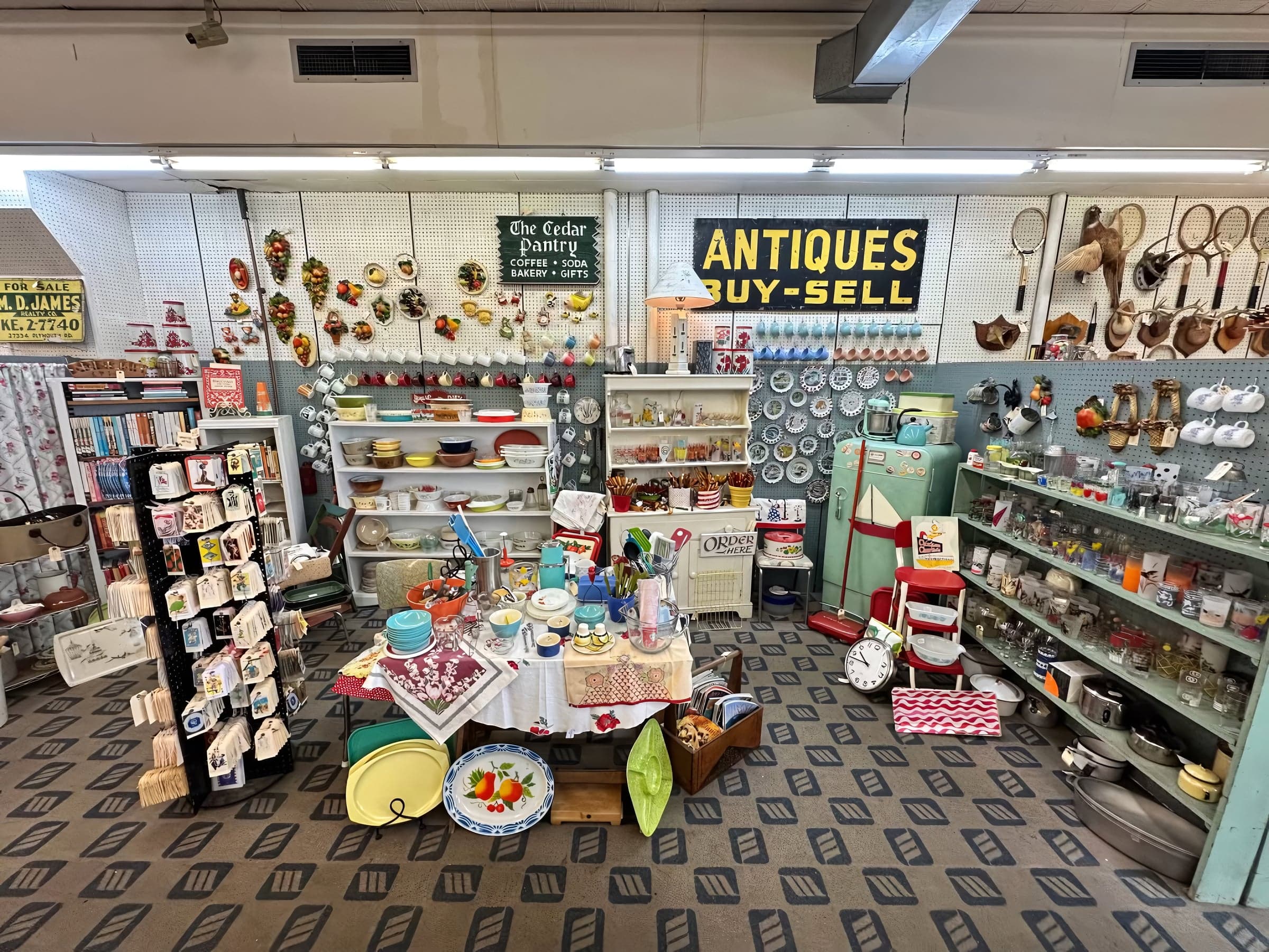 Wide view down an aisle of the mall with overhead signs reading 'The Cedar Pantry' and 'ANTIQUES BUY-SELL' above shelves of vintage kitchenware and glassware.
