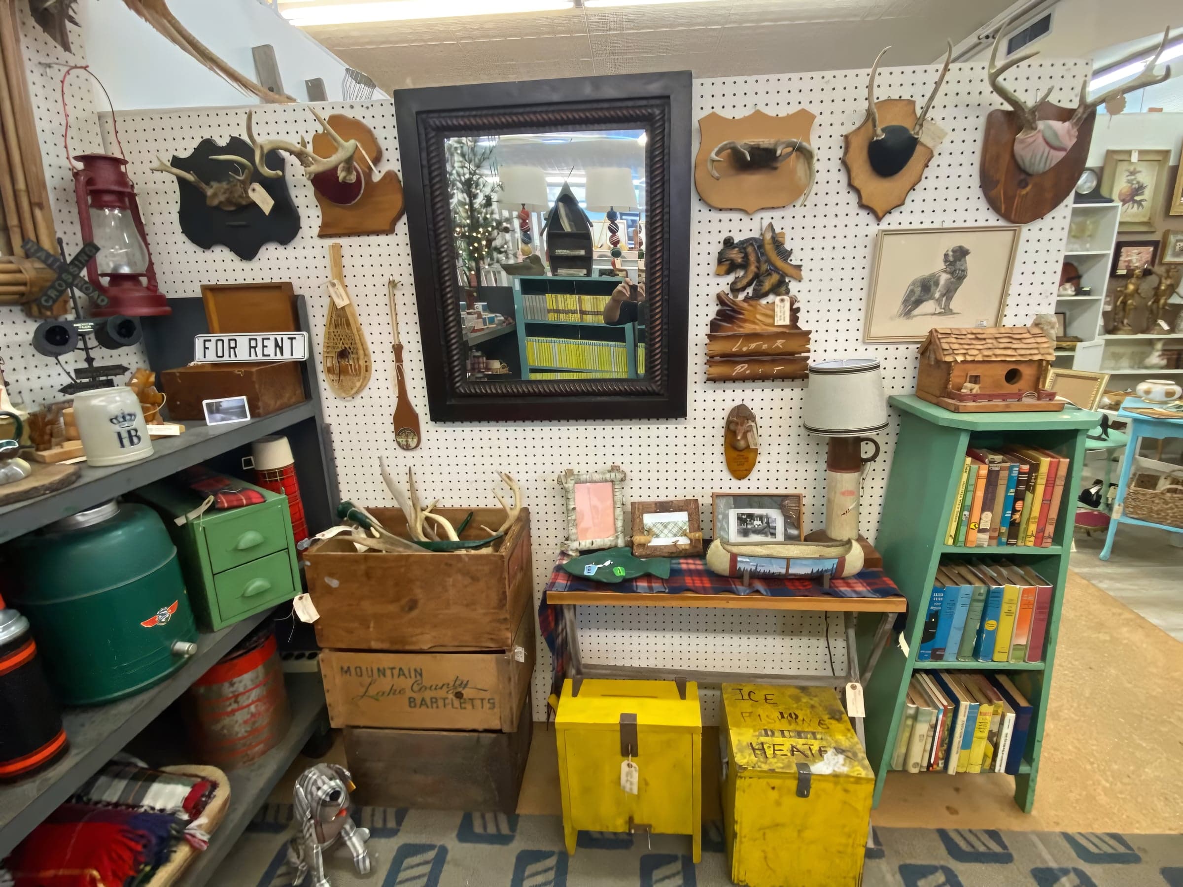 A vintage cabin-style booth: a framed pegboard mirror, mounted antlers, a 'FOR RENT' sign, plaid thermoses on shelves, and a yellow steamer trunk.