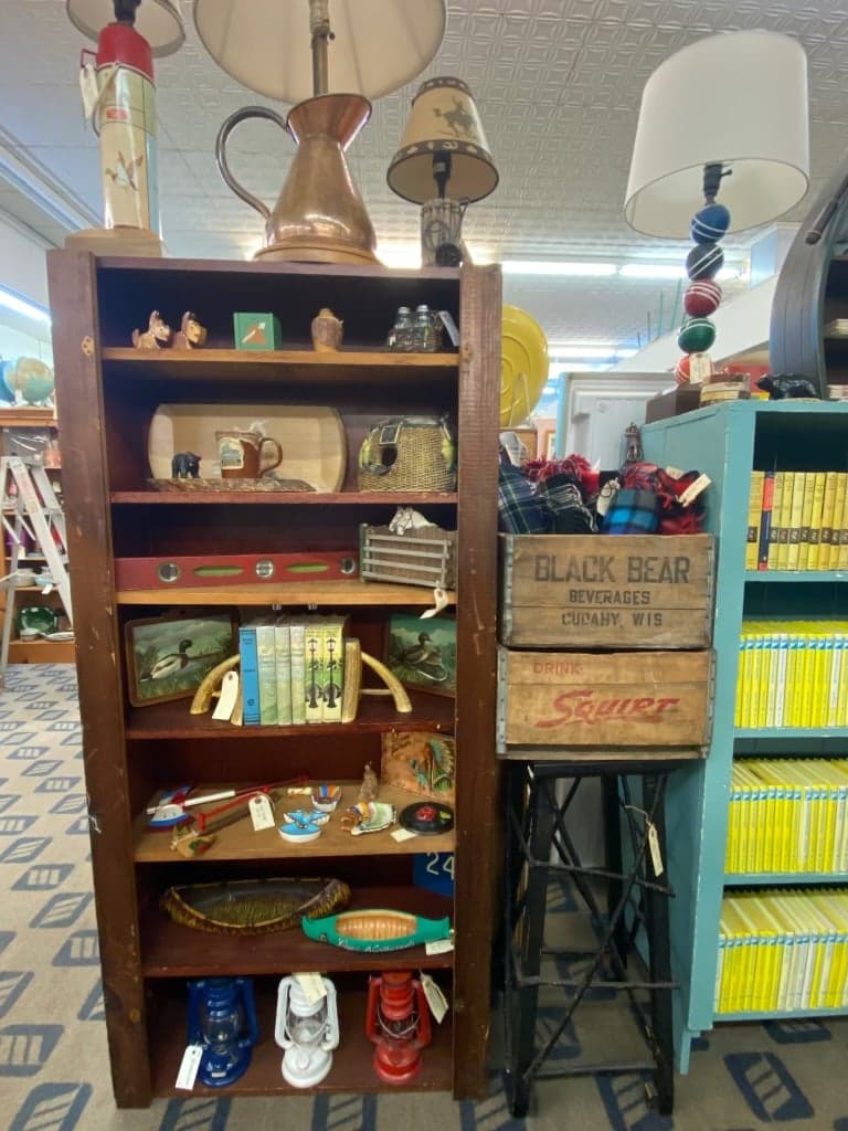 A tall wooden bookshelf inside one of the booths, holding vintage figurines, books, oil lanterns, and stacked Black Bear and Squirt soda crates.