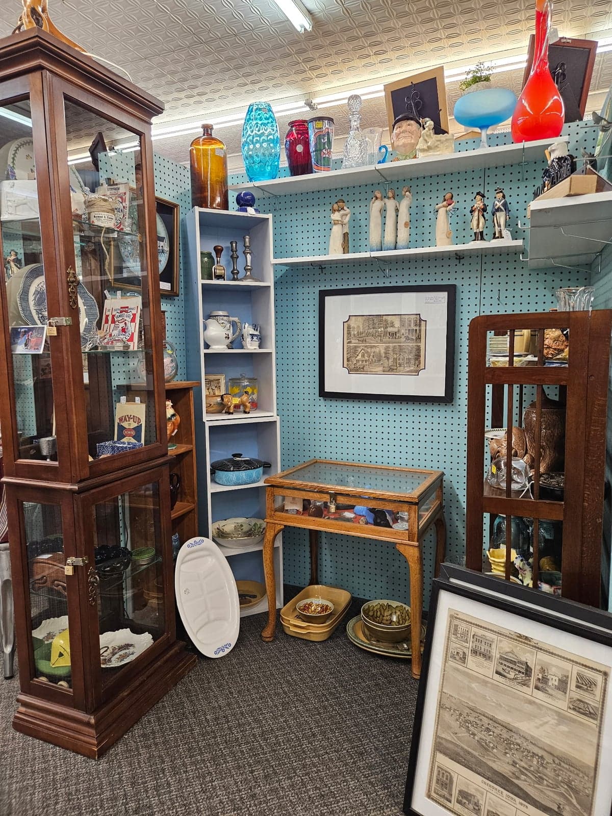 A dark wood curio cabinet of glassware beside shelves of vintage colored glass bottles, against a teal pegboard wall.